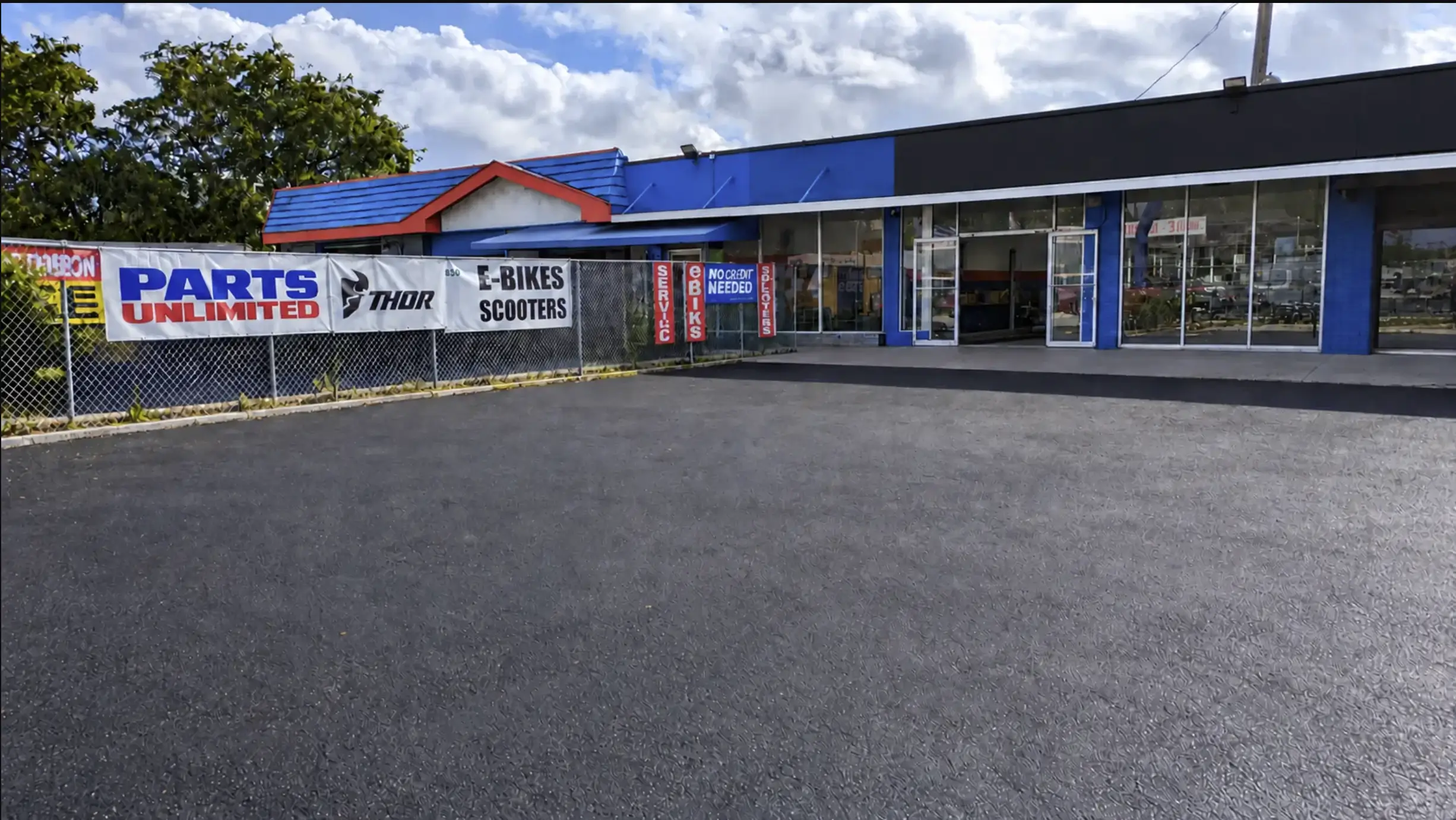 Front of 830 NE 79th St, Miami: a freshly paved front lot facing a blue-trimmed commercial building with glass storefronts and fenced frontage.